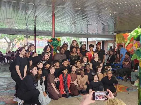 Large group of students in costumes pose together under a covered outdoor area with audience nearby.