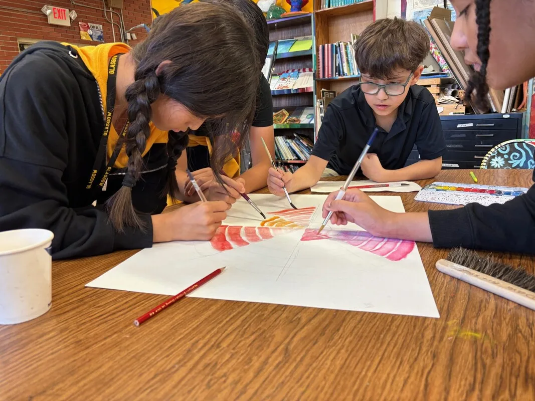 Four students collaborate on painting a colorful artwork together at a classroom table.