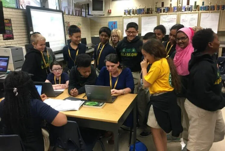Principal Erin Howard works with students gathered around laptops in the library. 