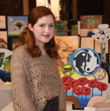 Student standing beside a display of colorful painted artwork and crafts on tables indoors.