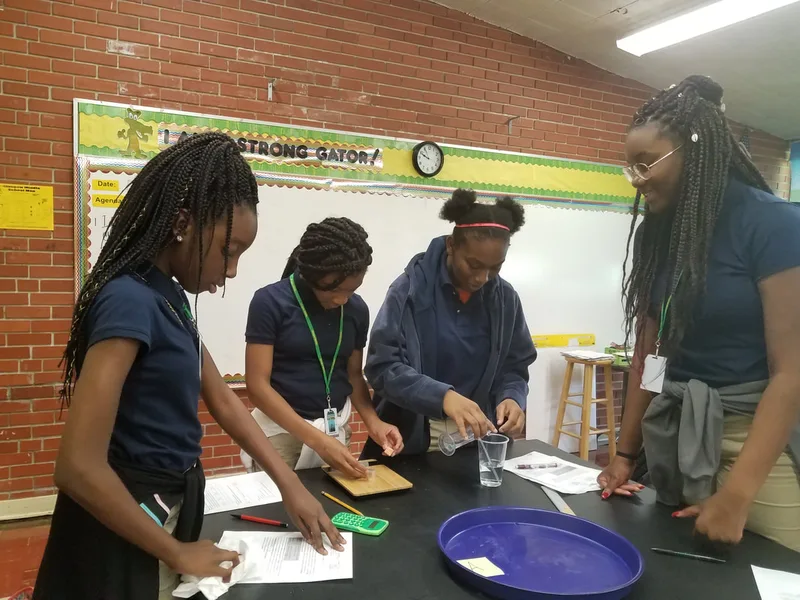 Four students conduct a science experiment with cups and tools on a classroom table.