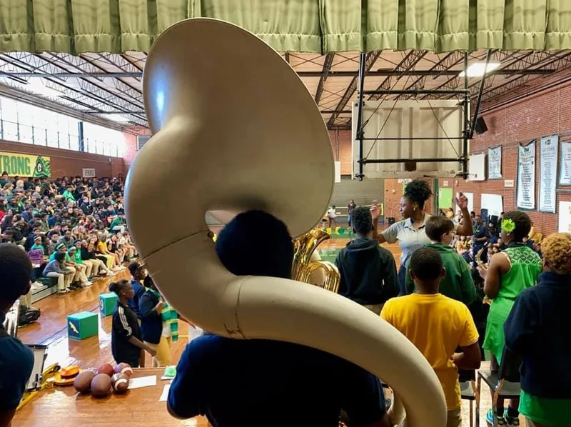 School gym assembly with students, a large sousaphone in foreground, and a crowd in bleachers.