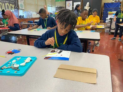 Student uses tools and glue to create a craft at a classroom table with classmates working nearby.