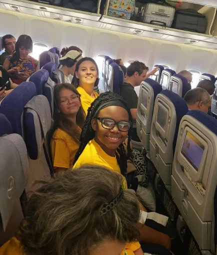 Students and teacher in yellow shirts sit on a plane, smiling toward the camera.
