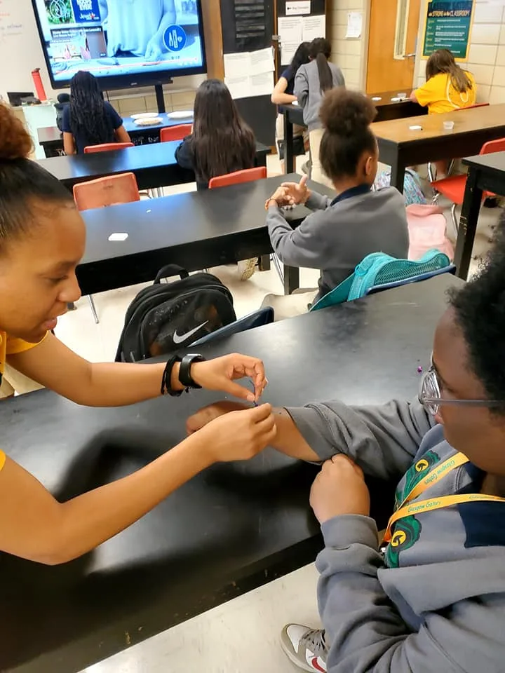 A student helps a student put on a bracelet. 