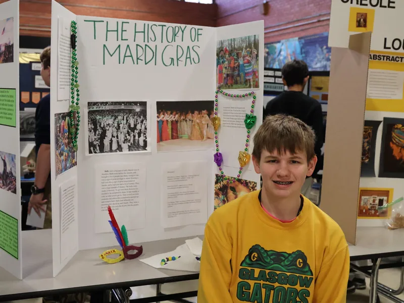Student sitting in front of a Mardi Gras history project board with photos, beads, and decorations.
