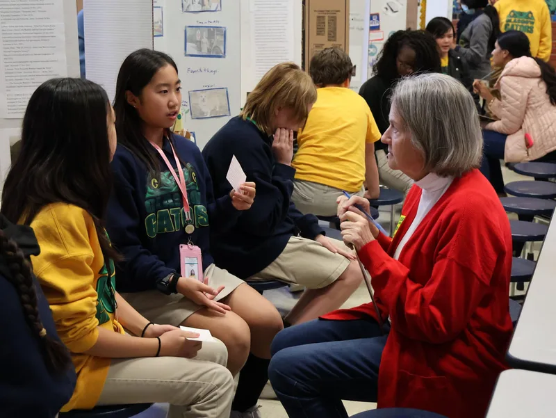 Students discussing a project with an adult evaluator seated in a room with display boards.

