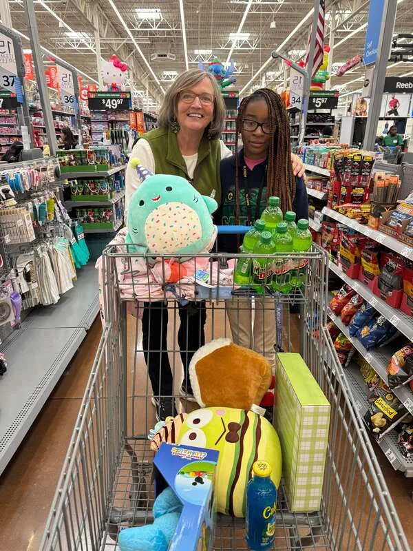 Adult and child with a cart of toys and drinks posing in a store aisle with holiday decor.
