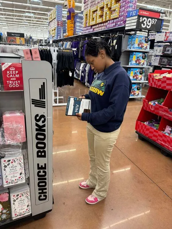 Person reading a book in a store aisle near a "Choice Books" display and boxed merchandise shelves.
