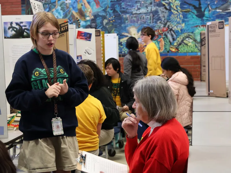 Student presenting a project to an adult evaluator with other displays in background.
