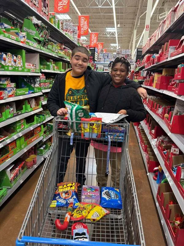 Two students posing with a shopping cart full of snacks in a store aisle decorated with holiday items.
