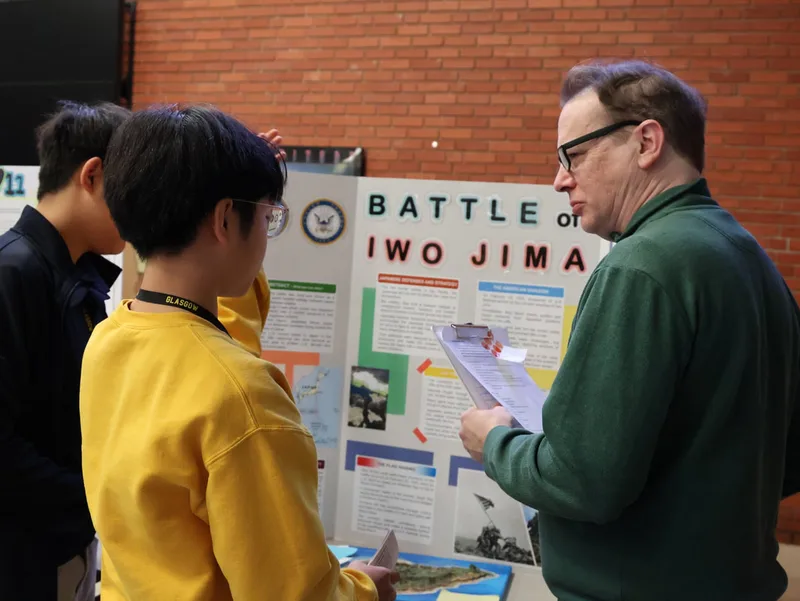 Student presenting Battle of Iwo Jima project to an adult with clipboard in a classroom.
