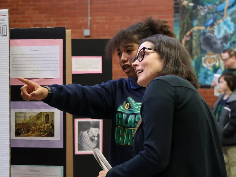 Student pointing at a display board while talking with an adult at a school project presentation.