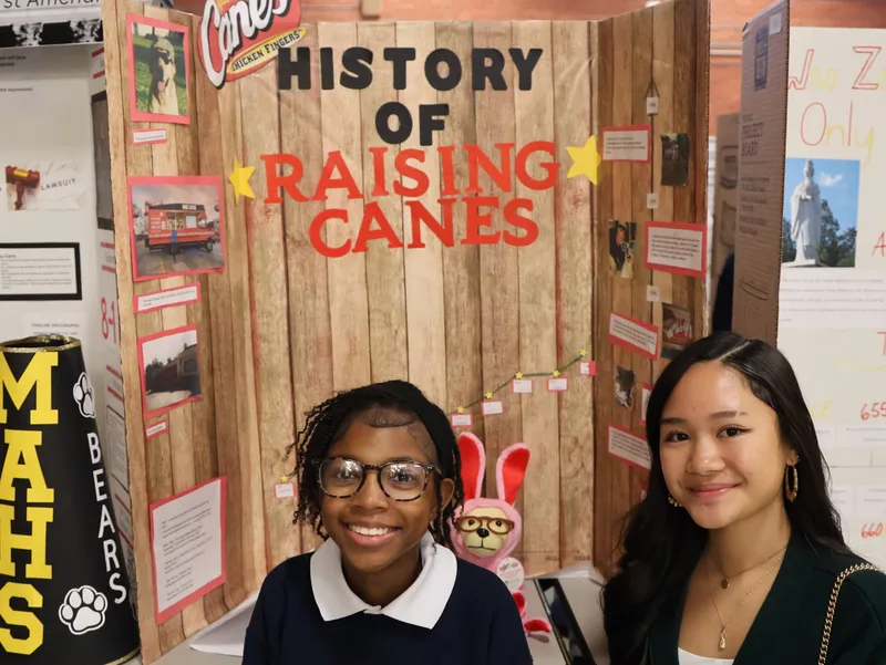 Two students posing by a “History of Raising Cane’s” display board with photos and timeline.

