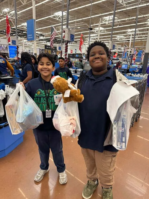 Two students at checkout holding shopping bags and a plush toy in a busy store.

