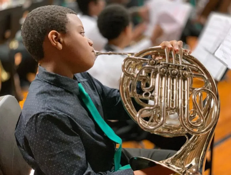 Student playing a French horn in a school band, reading sheet music during a performance. 