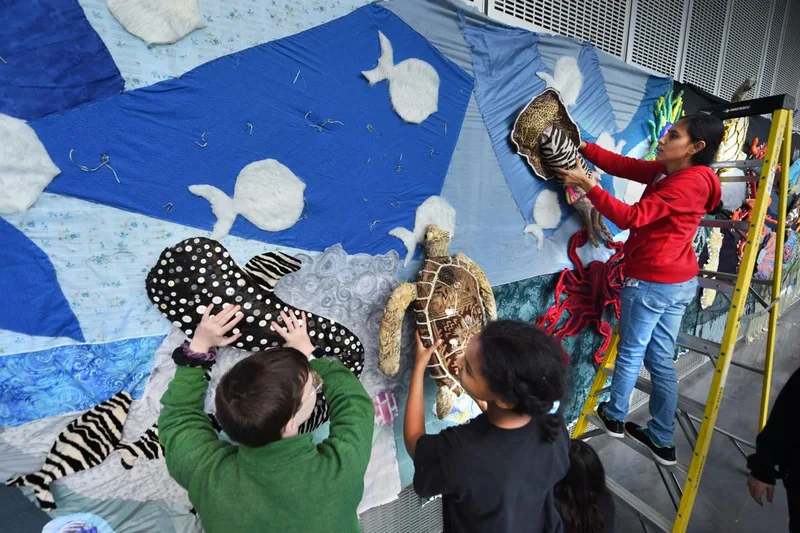 Students and an adult attach fabric sea turtles to a large ocean-themed wall display.