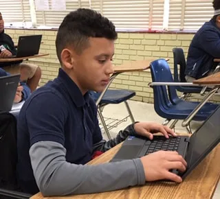 Student using a laptop at a desk in a classroom, focused on typing with classmates nearby.