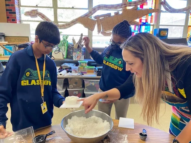 Students and teacher measure and mix materials in a bowl during a hands-on classroom activity.