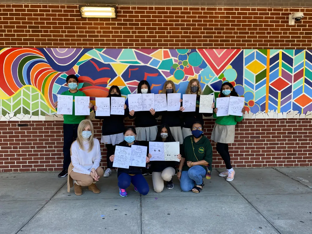 Students and adult hold drawings in front of a colorful mural on a brick wall.