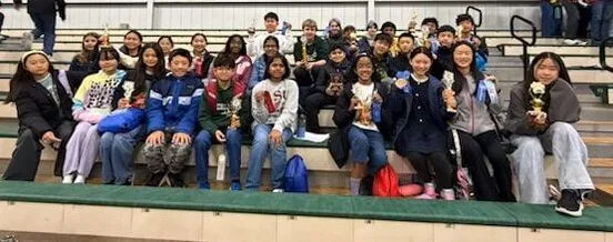 Large group of students seated on gym bleachers holding trophies and awards together.