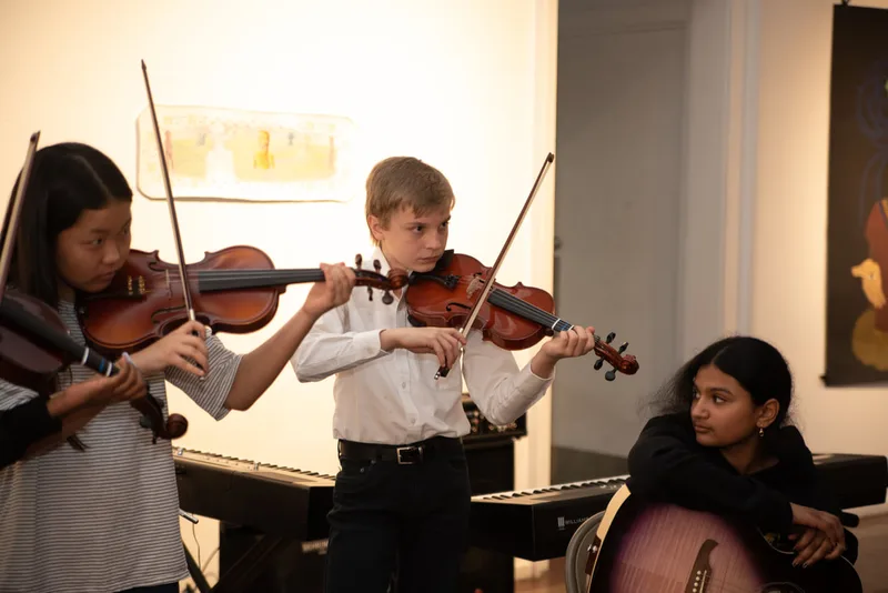 Two students play violins while another sits nearby with a guitar.