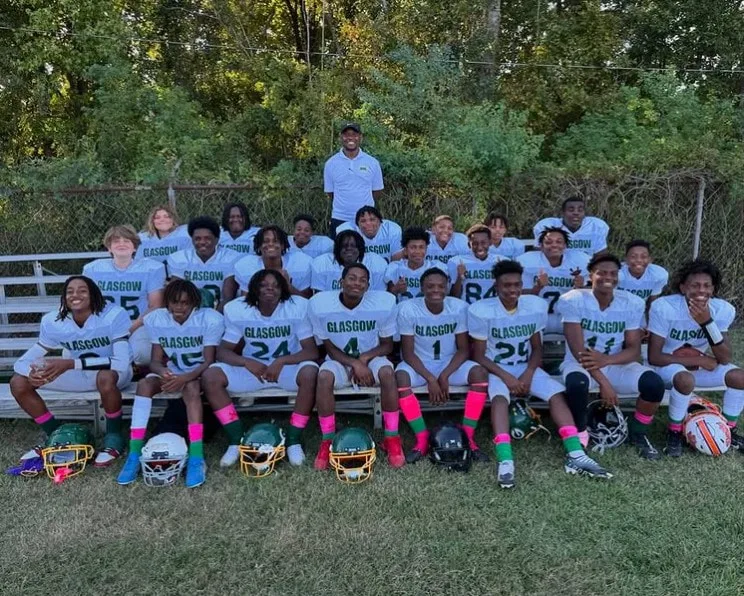 Football team in Glasgow jerseys sits on bleachers with coach standing behind on field.