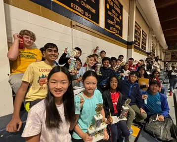 Group of students seated on bleachers holding trophies in a school gym with banners on wall.
