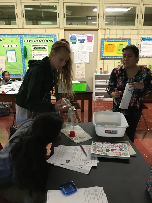 Two female students are measuring liquid in a cylinder. The teacher is standing near them. 