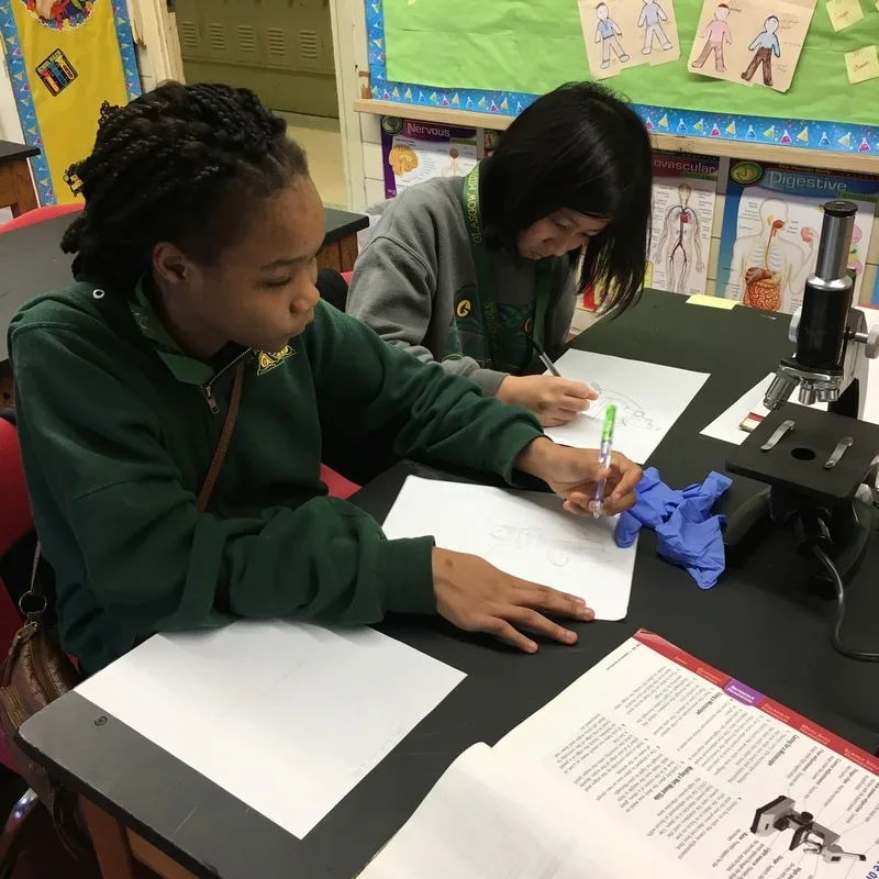 Two female students seated at a lab table. They are working on a science assignment. There is a microscope on the table. 