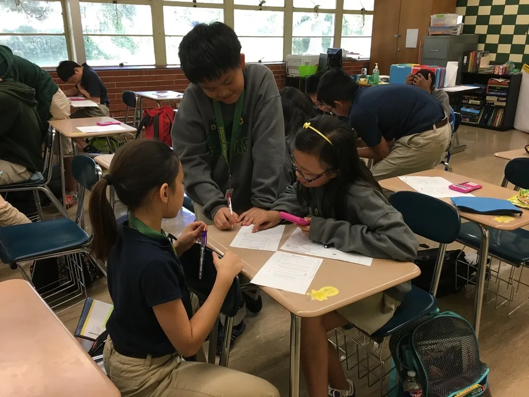 Two girls and a boy in a classroom working math problems. The girls are seated and the boy is standing next to them. 