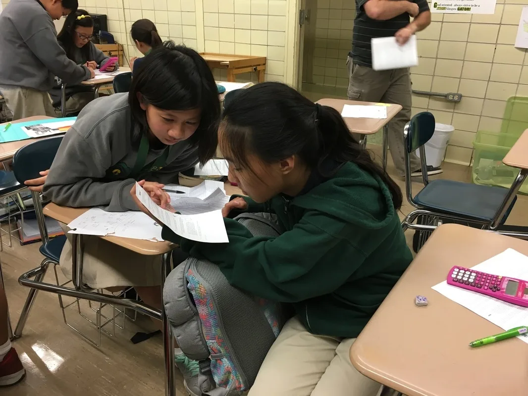 Two girls in a classroom working math problems. They are seated in desks. 