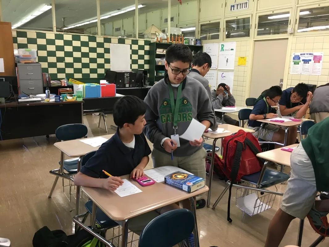 Two boys in a classroom working on math problems. One is seated at a desk and the other is standing next to him. 