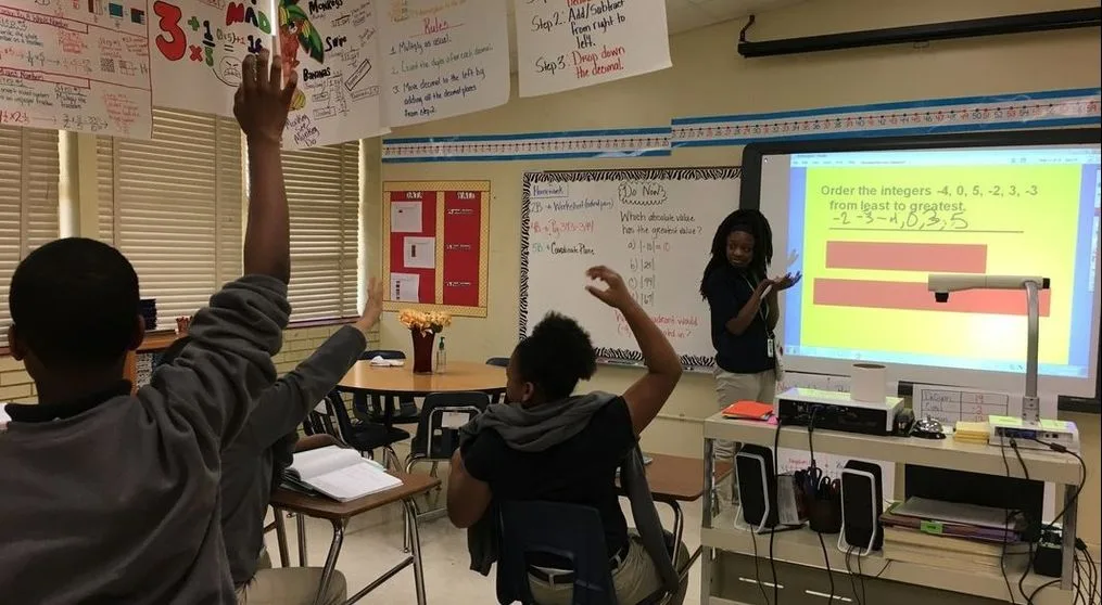 A classroom with a girl standing at a smartboard. She is explaining a math problem. Several students are raising their hands, waiting for her to call on them. 