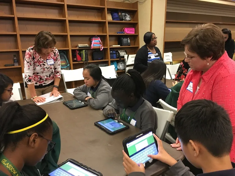 A group of students are seated at a table. Two volunteers are standing near them, assisting them. The students are working with tablets. 