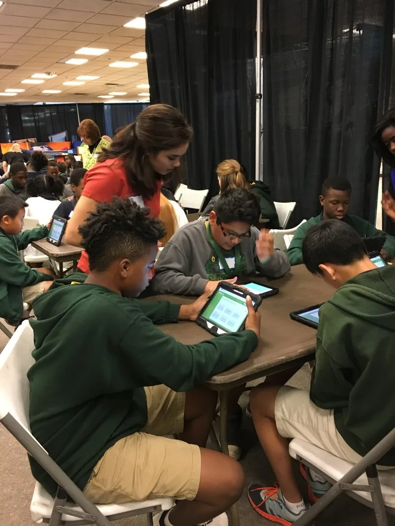 A group of students are seated at a table. A volunteer is standing near them, assisting them. The students are working with tablets. 