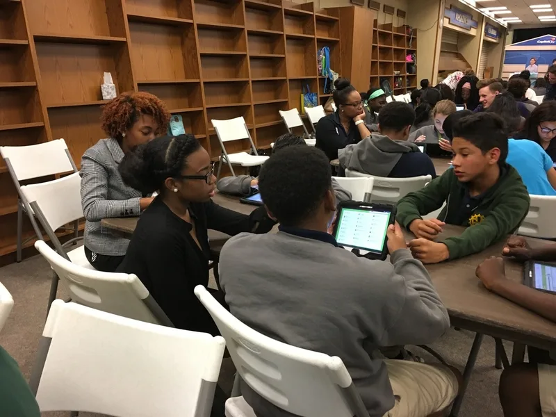 A group of students seated at a table with a volunteer. The students are working with tablets. 
