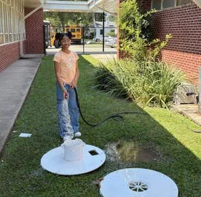 Student outdoors using a hose to spray water into a round ground cover near a school building.