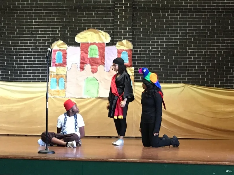 A boy dressed as Aladdin is seated on the ground. He is speaking to two students. 