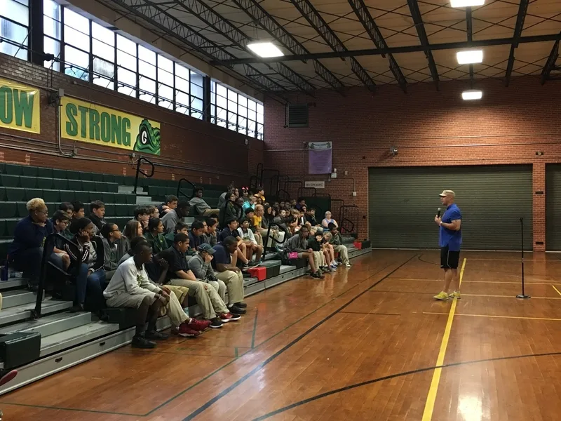 A group of students seated on bleachers in the gym. They are listening to a speaker. 
