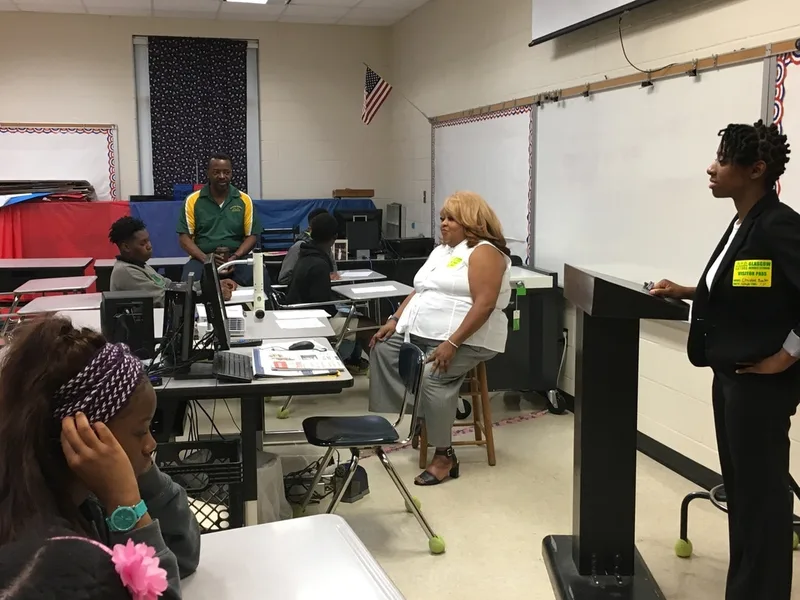 A group of students and a teacher are seated in a classroom. They are listening to two speakers. 
