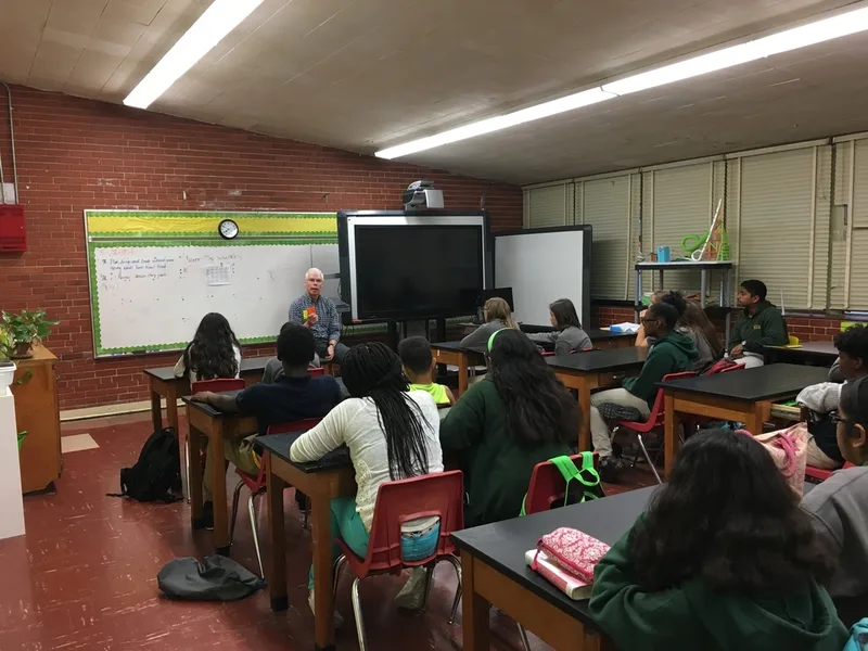 A group of students seated at tables in a classroom. They are listening to a speaker. 
