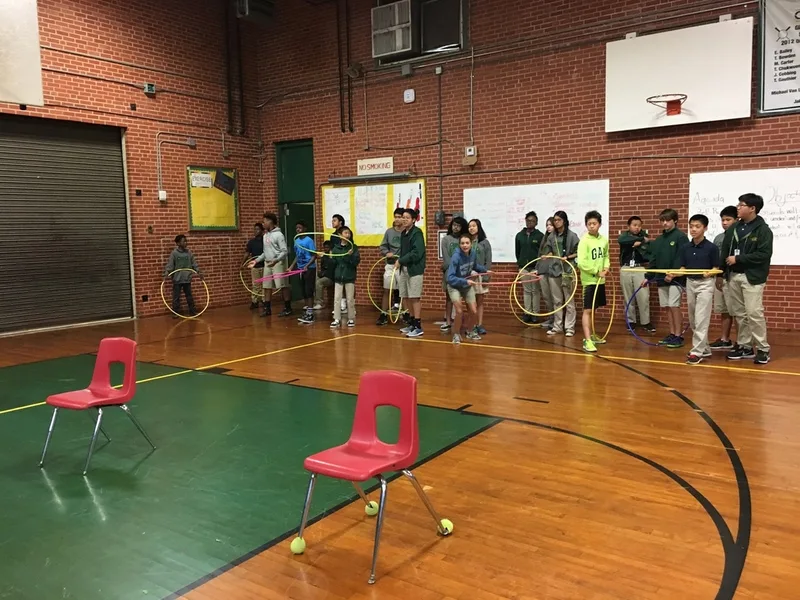 Students are lined up along the sidelines of the basketball court. Some are holding hula hoops. 
