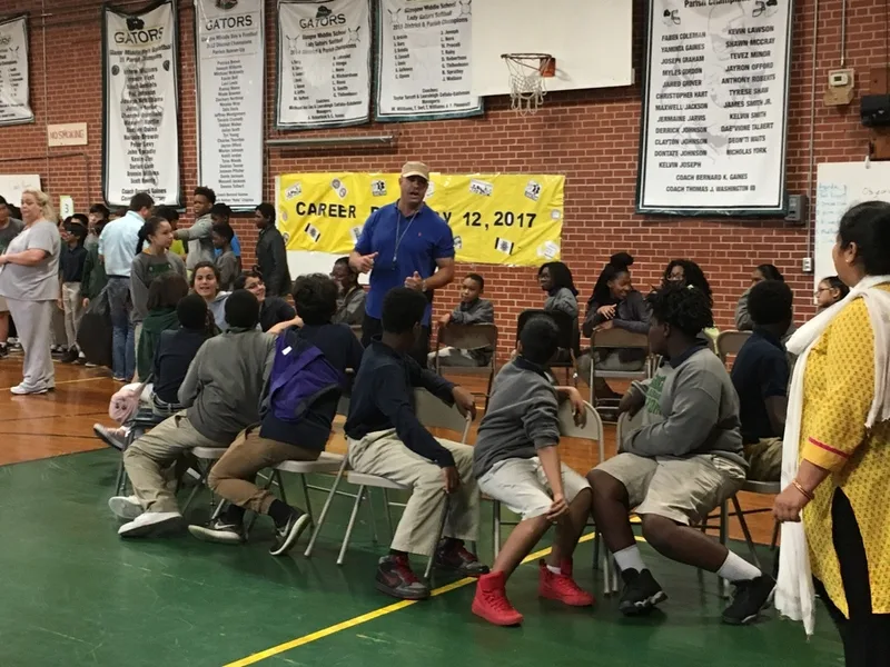 A group of students prepare to play musical chairs in the gym. A teacher is giving them instructions. 