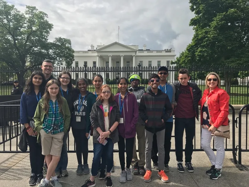 Group of students and adults pose in front of the White House behind a black fence.
