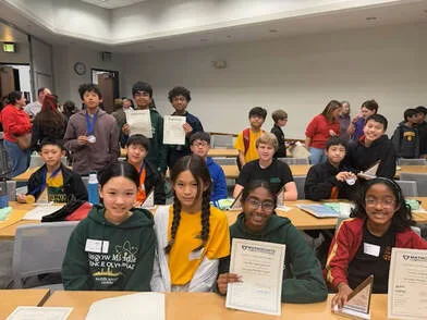 Group of students in a room holding certificates and awards, with peers and tables in view.