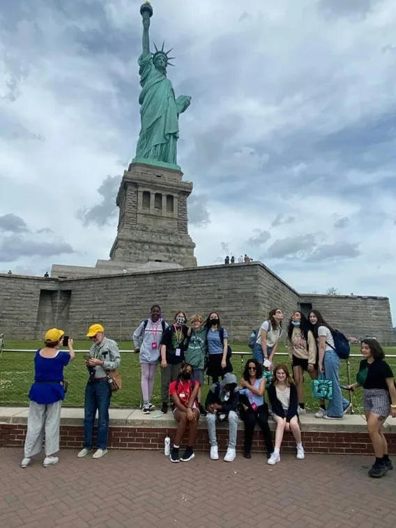 Group of students poses in front of the Statue of Liberty on a cloudy day during a trip.
