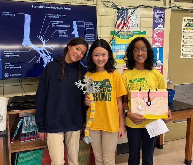 Three students display science models of lungs and skin in a classroom with diagram screen behind.