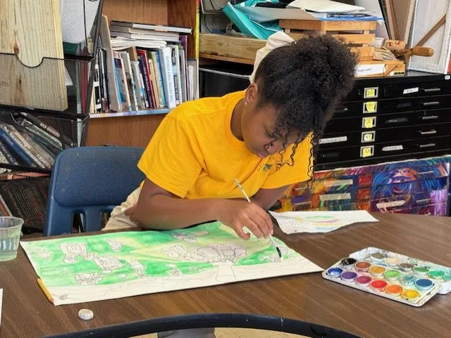 Student painting a detailed artwork on paper at a classroom table with watercolor set nearby.