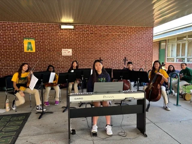 Student plays keyboard while classmates with string instruments sit behind music stands outdoors.